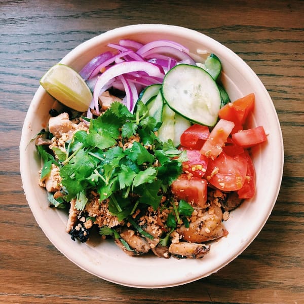A vibrant bowl of grilled chicken, fresh cilantro, red onions, cucumbers, tomatoes, and a lime wedge.