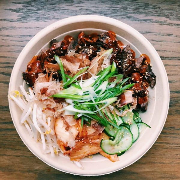 Asian-fusion bowl with beef, bean sprouts, kimchi, cucumbers, and green onions on a wooden table.