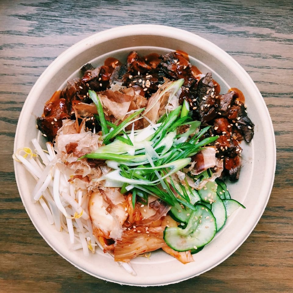 Asian-fusion bowl with beef, bean sprouts, kimchi, cucumbers, and green onions on a wooden table.